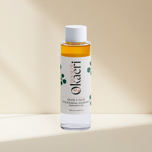 A bottle of Okaeri Aloe Calm Cleansing Essence with a white cap and white label, showing the amber liquid inside, set against a warm beige background with soft shadows.