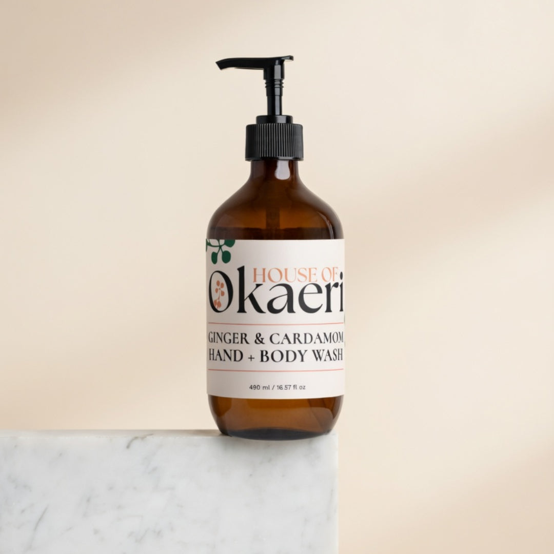 An amber glass pump bottle of House of Okaeri Ginger & Cardamom Hand & Body Wash, standing on a white marble surface against a warm beige background.