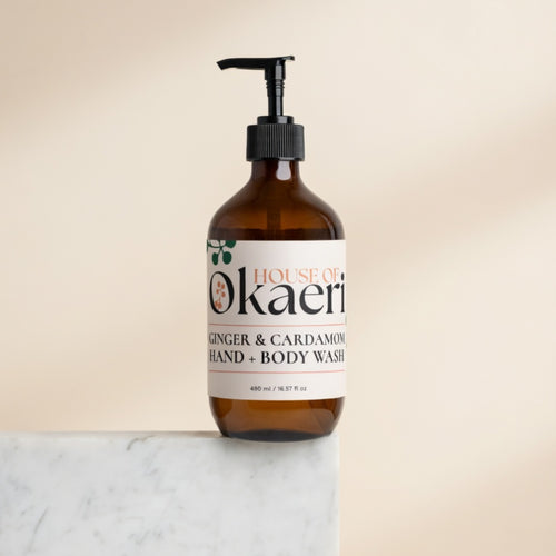 An amber glass pump bottle of House of Okaeri Ginger & Cardamom Hand & Body Wash, standing on a white marble surface against a warm beige background.