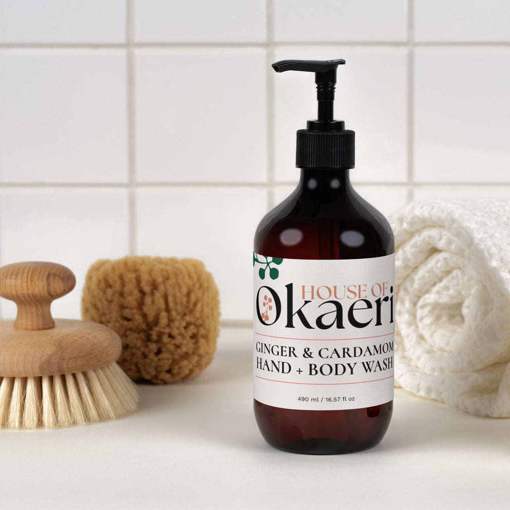A bottle of House of Okaeri Ginger & Cardamom Hand & Body Wash stands on a white surface against a white tiled wall, styled with a wooden brush, sponge, rolled towel, and bar of soap.