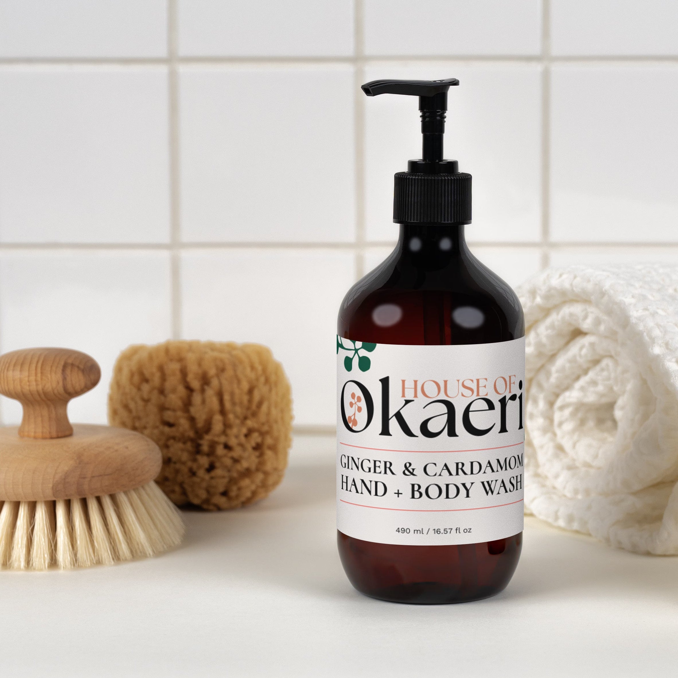 A bottle of House of Okaeri Ginger & Cardamom Hand & Body Wash stands on a white surface against a white tiled wall, styled with a wooden brush, sponge, rolled towel, and bar of soap.