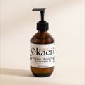 An amber glass pump bottle of House of Okaeri Ginkgo + Rosehip Body Serum, featuring a white label with black and orange text, set against a warm beige background.