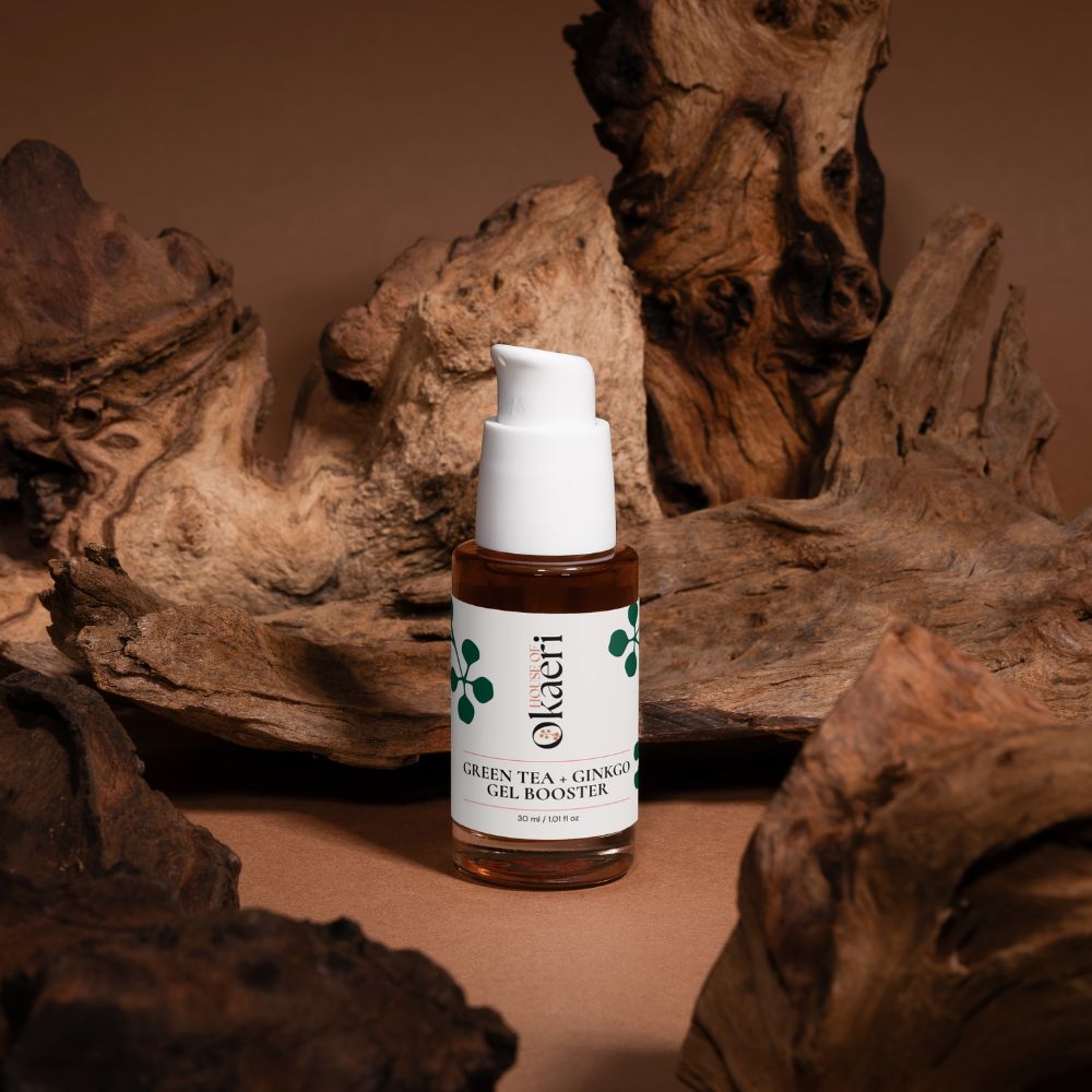 A bottle of Okaeri Green Tea + Ginkgo Gel Booster centered against a rustic background of driftwood logs on a warm brown surface.