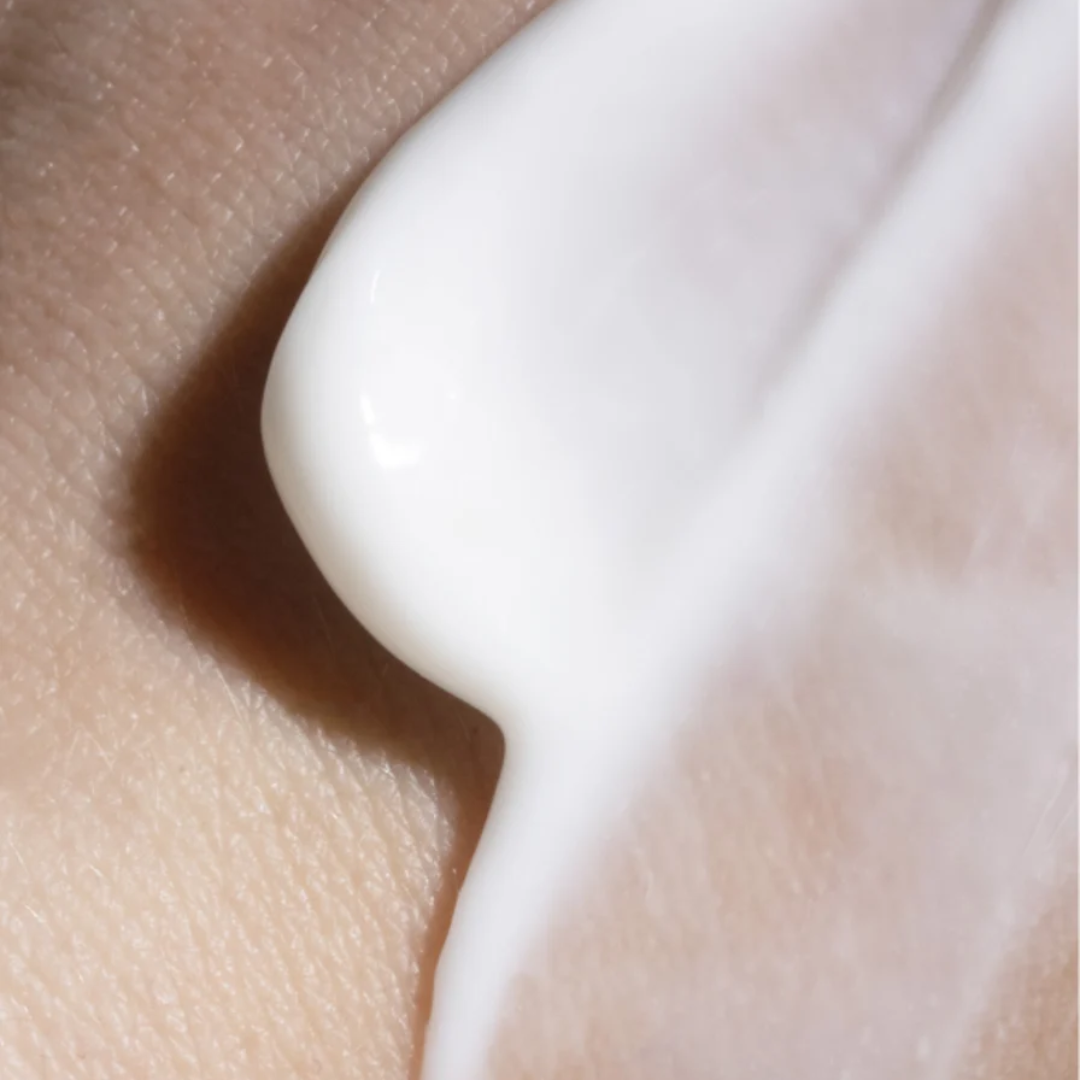 A close-up of a thick, creamy white lotion or serum being applied onto skin, showing a smooth, glossy texture against a warm beige skin tone.