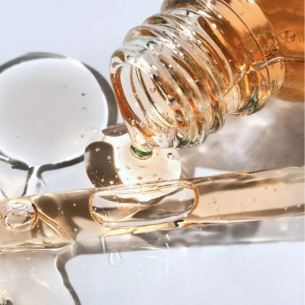 A close-up of a transparent bottle pouring a golden amber liquid over a glass surface, with droplets and bubbles visible against a soft light background.
