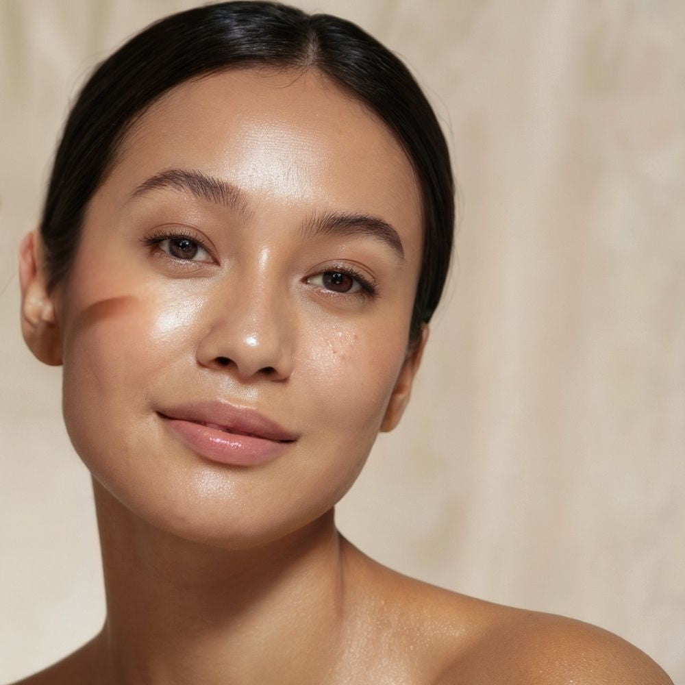 A close-up portrait of a woman with a warm, radiant complexion, wearing a light brown contour makeup sweep on her cheek, set against a soft beige textured background.